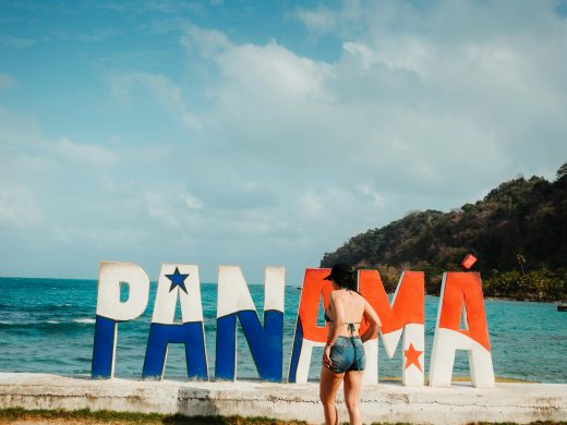 Woman in shorts admires colorful 'Panamá' sign by the sea on a sunny summer day.